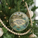 A round ornament hanging on a Christmas tree, featuring an illustration of a tropical beach scene with palm trees, a lighthouse, and a bird.