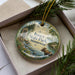 A decorative ornament in a box, featuring a moose and a scenic landscape with a river and pine trees.