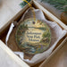 A decorative ornament in a box, featuring a scenic landscape with a bird, a cabin, and a river.