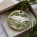 A decorative ornament in a box, featuring a scenic landscape with a waterfall and greenery.