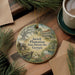A round ornament with a painting of a plantation house and a pond, surrounded by pine branches and a cup of coffee on a wooden surface.
