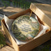 A decorative ornament in a wooden box, featuring a scenic landscape with a house, trees, and flowers.