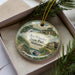 A decorative ornament in a box, featuring a scenic landscape with a house, trees, and a body of water.