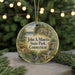 A round ornament with a painting of a scenic landscape, featuring a river, bridge, and trees, hangs in front of a Christmas tree with lights.