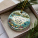 A decorative ornament in a box, featuring an illustration of a tropical beach scene with palm trees, coral reefs, and a lighthouse.