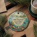 A decorative ornament featuring a tropical scene with a lighthouse, palm trees, and a coral reef, placed on a wooden surface alongside a cup of coffee and some pine branches.