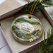 A decorative ornament in a box, featuring a scenic landscape with a bridge, water, and flowers.