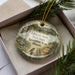 A decorative ornament in a box, featuring a scenic landscape with a bridge, flowers, and a bird.
