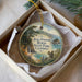 A round ornament with a painting of a forest scene and a squirrel, placed in a white box with a gold string.