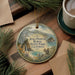 A round ornament with a scenic landscape of Julia Pfeiffer Burns State Park, California, featuring a squirrel and pine trees, is displayed on a wooden surface alongside a cup of coffee and some papers.