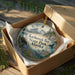 A circular ornament in a wooden box, featuring a scenic landscape with mountains, water, and wildlife, including an eagle and a whale.
