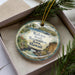 A circular ornament with a tropical beach scene and palm trees, placed in a box with pine branches.