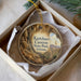 A circular ornament with a painting of a canyon and a plant, placed in a box with a white tissue paper lining.