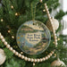A circular ornament hanging on a Christmas tree, featuring a scenic landscape with a lake, mountains, and a bird.