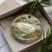 A decorative ornament in a box, featuring a scenic landscape with a body of water, trees, and a city skyline in the background.