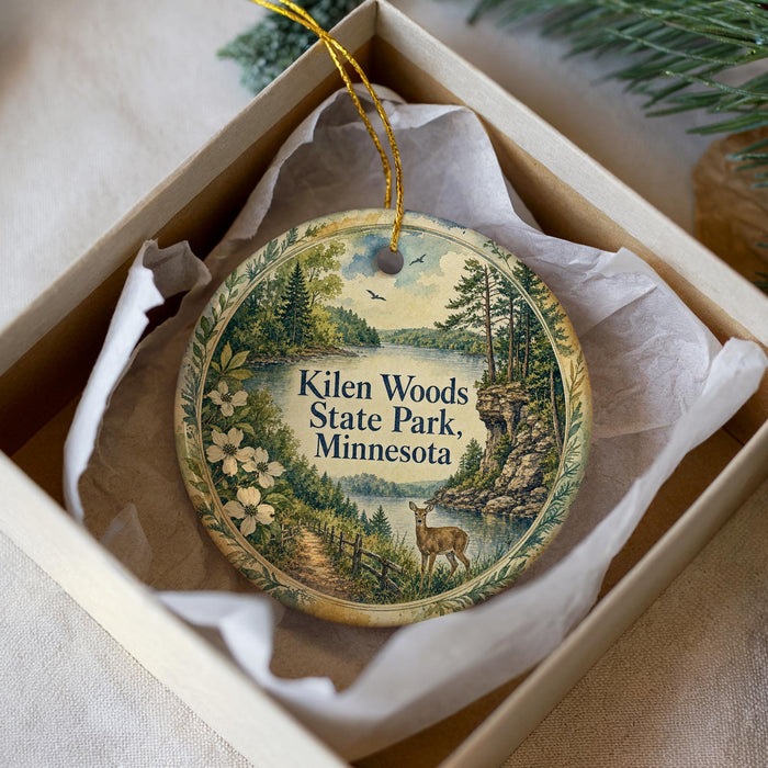 A decorative ornament in a box, featuring a scenic landscape with a lake, trees, and a deer.