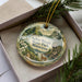 A decorative ornament in a box, featuring a scenic landscape with a deer and pine trees, along with the text "Kilen Woods State Park, Minnesota".