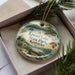 A decorative ornament in a box, featuring a scenic landscape with a lake, cabin, and pine trees.