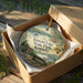 A decorative ornament in a box, featuring a scenic landscape with a bench, trees, and a body of water.