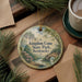 A decorative ornament with a scenic landscape of Kentucky State Park, featuring a river, trees, and a bridge, is displayed on a wooden surface alongside a cup of coffee and some pine branches.