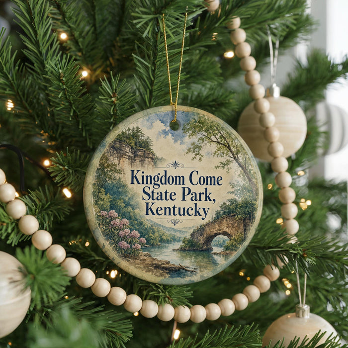 A round ornament hanging on a Christmas tree, featuring a scenic landscape with a river and trees.