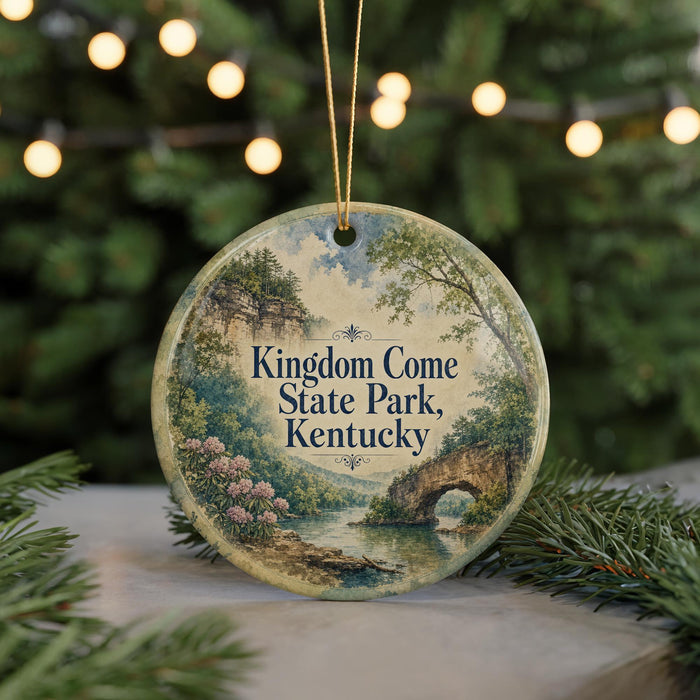 A round ornament with a scenic landscape of Kentucky State Park, featuring a bridge, trees, and a river, is displayed against a blurred background of Christmas lights.