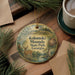 A decorative ornament featuring a deer and a landscape scene, with the text "Kolomoki Mounds State Park, Georgia" printed on it. The ornament is placed on a wooden surface, surrounded by pine branches and a cup of coffee.