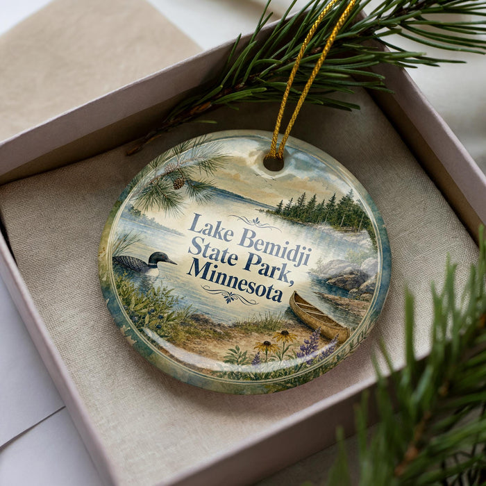 A decorative ornament with a lake and forest scene, featuring a bird and pine trees, is displayed in a box with pine branches.