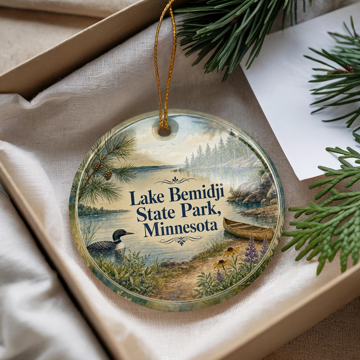 A decorative ornament with a lake scene and pine trees, featuring a bird and a canoe, is displayed in a box.