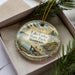 A decorative ornament in a box, featuring a scenic landscape with a lake, dock, and pine trees.