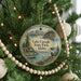 A round ornament hanging on a Christmas tree, featuring a scenic landscape with mountains, a lake, and a cabin.