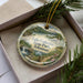 A decorative ornament in a box, featuring a scenic landscape with a waterfall and greenery, with the text "Lake Catherine State Park, Arkansas" printed on it.