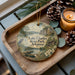 A decorative ornament featuring a scenic landscape of Lake Charles State Park, Arkansas, with a bird, a cabin, and a lake. The ornament is displayed on a wooden tray, accompanied by pine cones and a lit candle.