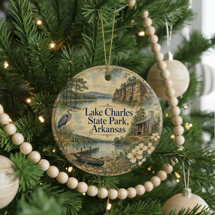 A round ornament hanging on a Christmas tree, featuring a scenic landscape with a lake, trees, and a cabin.