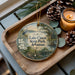 A decorative ornament featuring Lake Chico State Park, Arkansas, with a bird and a bridge in the background, placed on a wooden tray alongside pine cones and a lit candle.