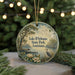A round ornament with a scenic landscape featuring a lake, trees, and birds, hanging in front of a Christmas tree with lights.
