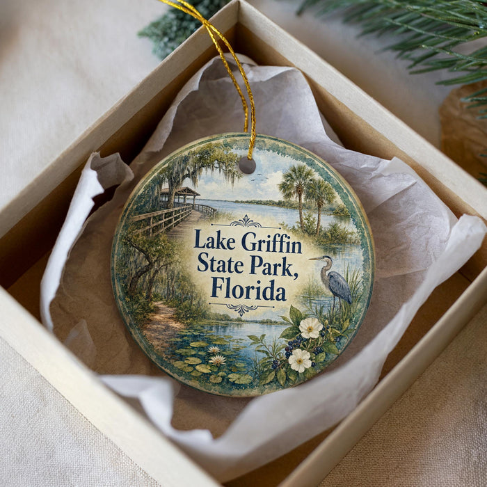 A decorative ornament in a box, featuring a scenic landscape with a lake, palm trees, and a bird, along with the text "Lake Griffin State Park, Florida".