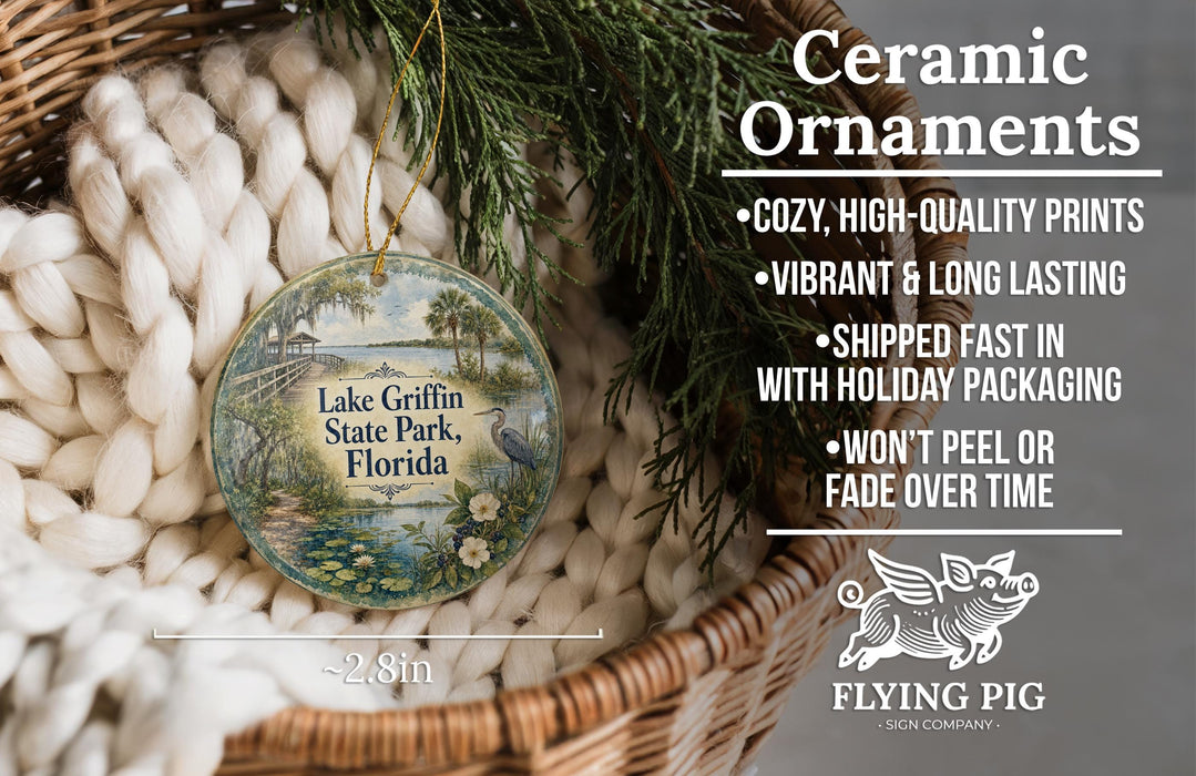 Ceramic ornaments, featuring a round ornament with a landscape design, are displayed in a basket with greenery.
