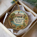 A decorative ornament in a box, featuring a scenic landscape with a lake, mountains, palm trees, and a bird.