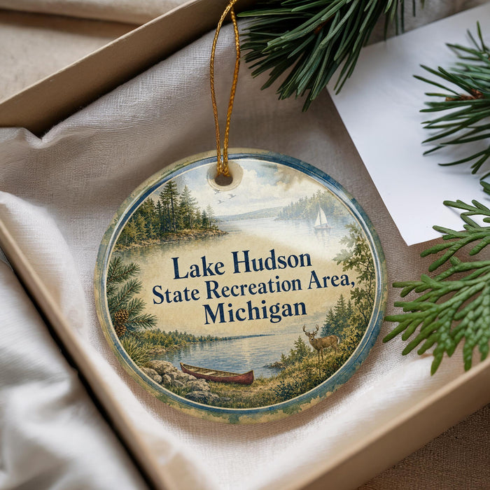 A round ornament with a scenic lake and forest landscape, featuring a canoe and a deer, is displayed in a box with pine branches.