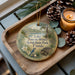 A decorative ornament with a bird and flowers, placed on a wooden tray next to pine cones and a lit candle.