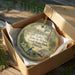 A round ornament in a box, featuring a bird and a landscape scene, with text that reads "Lake June-in-Winter Scrub State Park, Florida".