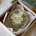 A decorative ornament in a box, featuring a scenic landscape with a lake, trees, and a path leading to a dock.