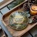 A wooden tray displaying a decorative ornament with a scenic lake landscape, pine cones, and a lit candle.