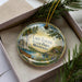 A decorative ornament in a box, featuring a scenic landscape with a lake, pine trees, and a canoe, along with the text "Lake Louise State Park, Minnesota".