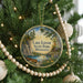A circular ornament hanging on a Christmas tree, featuring a scenic landscape with a lake, pine trees, and a cabin.