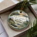 A decorative ornament with a lake and surrounding scenery, featuring a boat and a bird, is displayed in a box with pine branches.
