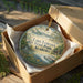 A decorative ornament in a box, featuring a scenic landscape with a lake, trees, and a boat, with the text "Lake Lurleen State Park, Alabama" printed on it.