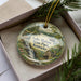 A decorative ornament in a box, featuring a scenic landscape with a waterfall and a house, with the text "Lake Malone State Park, Kentucky" printed on it.