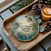 A decorative ornament with a scenic image of Lake Manatee State Park, Florida, featuring a bird, palm trees, and a body of water. The ornament is placed on a wooden tray, with pine cones and a lit candle nearby.